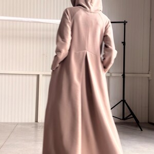 May include: A long, hooded coat in a soft, muted pink color. The coat features a flowing design with a pleated back and long sleeves. The model is facing away from the camera, showcasing the coat's silhouette.