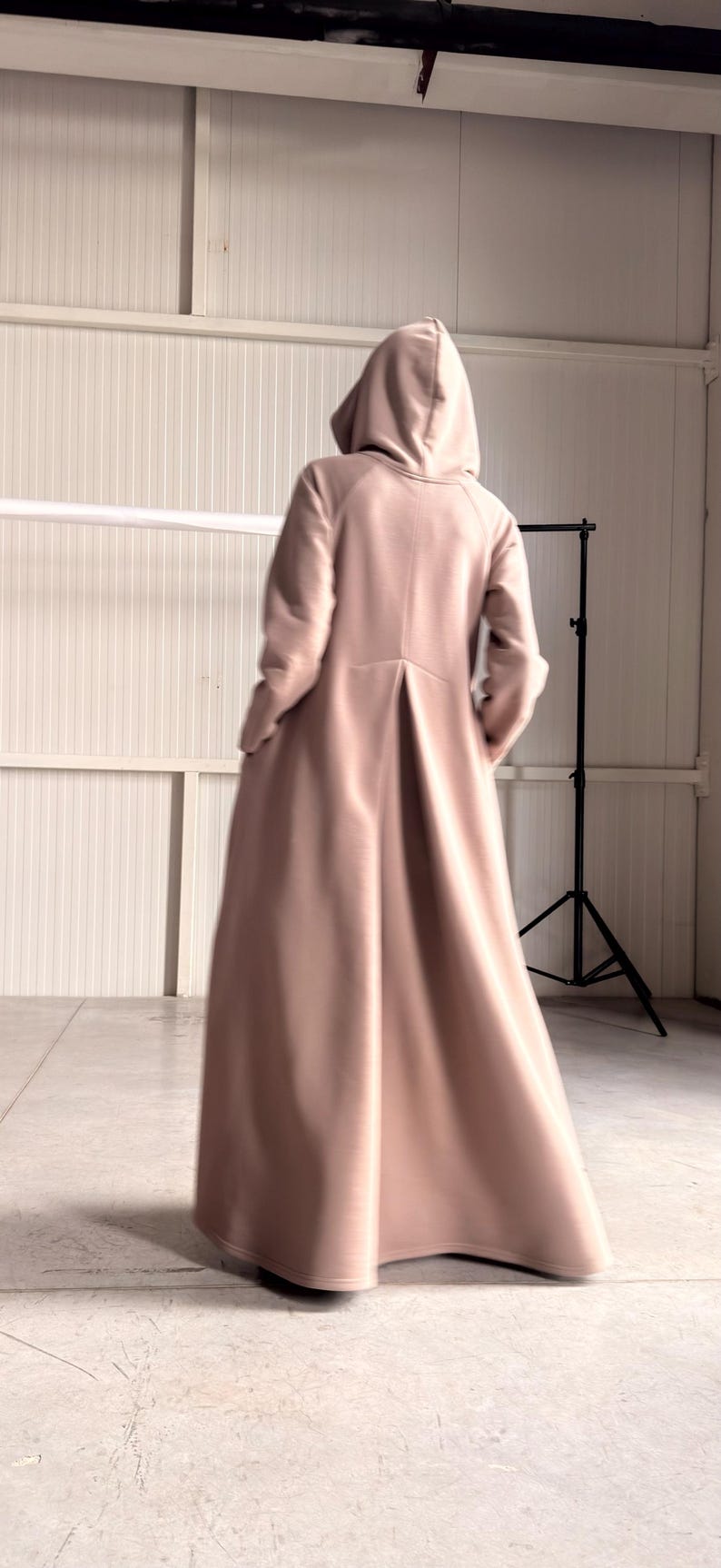 May include: A long, hooded coat in a soft, muted pink color. The coat features a flowing design with a pleated back and long sleeves. The model is facing away from the camera, showcasing the coat's silhouette.