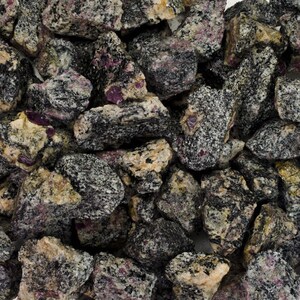 May include: A close-up of a pile of rough, dark gray and black stones with flecks of pink and white. The stones are irregularly shaped and have a rough, textured surface.