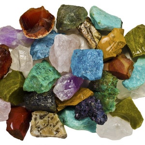 May include: A collection of rough, unpolished gemstones in various colors, including green, blue, purple, red, and white. The stones are arranged in a pile on a white background.
