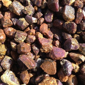 May include: A close-up view of a pile of rough, unpolished ruby gemstones. The rubies are a deep red color with a hint of purple. They are various sizes and shapes.