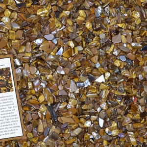 May include: A close-up of a pile of tiger's eye stones. The stones are a mix of brown, gold, and black. The stones are small and irregular in shape. The image includes a card with the text "Tiger Eye Home Decor/Healing Stones".