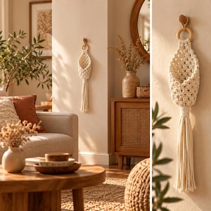 Op de afbeelding: Een crèmekleurige macramé wandmand met een gevlochten ruitpatroon, een ronde houten ring aan de bovenkant voor bevestiging en een lange, dikke franje-kwast die onder de buidel hangt, opgehangen aan een houten wandhaak.