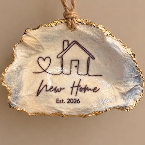 May include: A decorative oyster shell ornament with a gold-painted edge and a jute twine hanger. The surface features a black line drawing of a heart and a house, with the text New Home and Est. 2026 printed in a cursive and sans-serif font.