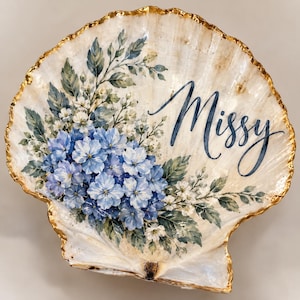 May include: A large, off-white scallop shell featuring a hand-painted watercolor illustration of blue hydrangea flowers and green leaves. The name Missy is written in blue cursive script on the right side, and the shell edge is trimmed in gold.