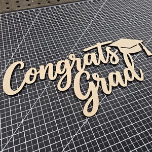 May include: A wooden graduation cake topper with the words "Congrats Grad" in a cursive font, along with a mortarboard. The topper is light brown and sits on a black cutting mat.