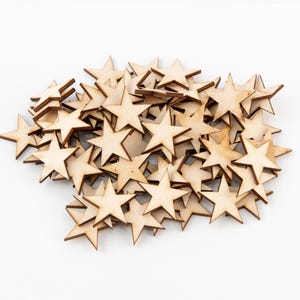 Puede incluir: Un mont&oacute;n de peque&ntilde;os recortes de madera en forma de estrella. Las estrellas son de color marr&oacute;n claro con un borde marr&oacute;n m&aacute;s oscuro y est&aacute;n esparcidas sobre un fondo blanco. Estos suministros de artesan&iacute;a son ideales para proyectos de bricolaje y decoraciones.