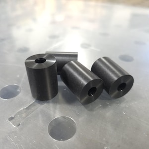 May include: Four small, cylindrical metal objects with a dark gray finish. Each cylinder has a hole drilled through the center. The objects are arranged on a reflective, metallic surface.