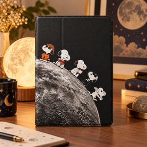 May include: A black tablet case featuring a graphic of Snoopy characters walking along the surface of the moon. The design includes multiple Snoopy figures in various poses against a textured lunar landscape set on a dark background.