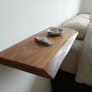 May include: A rectangular floating shelf made of natural wood with a live edge, mounted to a white wall. On the shelf sits a silver-toned wristwatch with a metal link band and a small, shallow, metallic bowl containing a single gold-colored ring.