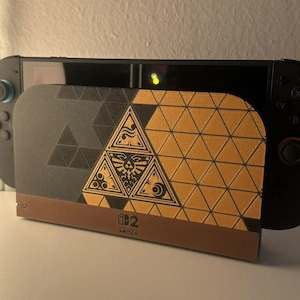 Puede incluir: Una consola Nintendo Switch negra acoplada en una base de carga personalizada con un patrón geométrico dorado y gris oscuro. El panel frontal muestra un emblema Triforce central con símbolos intrincados y el logo de Nintendo Switch abajo.