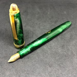 May include: A fountain pen with a marbled emerald green barrel and cap, featuring gold-toned metal accents including a clip, a rounded cap finial, and a metallic nib. The pen body displays a swirling green pattern with a gold-coloured logo imprint.