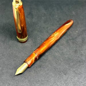 May include: A fountain pen with a marbled amber and brown resin body and a gold-coloured nib. The pen features a matching cap with a gold-toned clip and band. The barrel displays the engraved text "Admiral" in a gold script font near the centre.