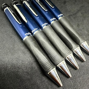 Op de afbeelding: Een set van vier donkerblauwe mechanische pennen met zwarte rubberen grepen en zilverkleurige metalen clips. Elke pen is voorzien van de tekst PhD in het wit op de houder, een zilverkleurige conische punt en een zwarte drukknop aan de bovenkant.