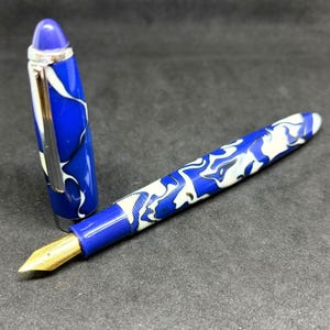 May include: A fountain pen with a marbled blue, white, and black resin body and cap. The pen features a gold-toned metal nib, a silver-toned metal clip on the cap, and a smooth, polished finish throughout the barrel and the removable cap.