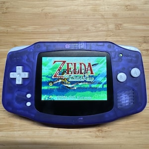 May include: A translucent purple handheld video game console with a white directional pad and A and B buttons. The screen displays the title The Legend of Zelda with the text FRAGILE HANDLE WITH CARE DO NOT FALL NOT PRESSURE Peel off this film before install.