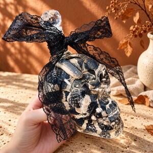 May include: A clear glass skull-shaped bottle featuring a black and white snake pattern inside the glass. The bottle is topped with a faceted clear glass stopper and is tied with a decorative black lace ribbon around the neck area.