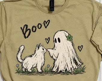 May include: A tan t-shirt featuring a black graphic print of a white cat and a ghost standing in grass. The ghost has a green four-leaf clover on its head. The text "Boo" is printed in a handwritten style above the figures, accompanied by three black hearts.