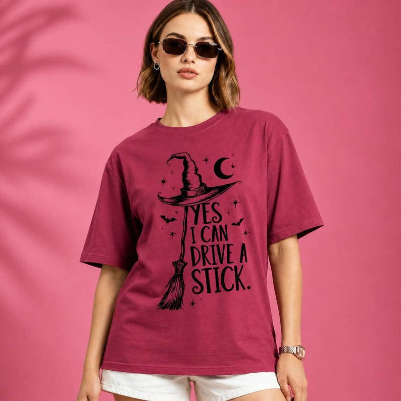 May include: A maroon short-sleeve t-shirt featuring a black graphic of a witch hat and a broomstick. The text reads, YES I CAN DRIVE A STICK, accompanied by small star, moon, and bat illustrations. The shirt has a crew neckline and a relaxed fit.