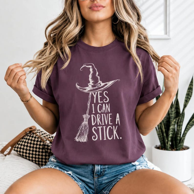 May include: A dark purple short-sleeve t-shirt featuring a white graphic of a witch hat perched atop a broomstick. The text printed in white reads, YES I CAN DRIVE A STICK. The shirt has a crew neckline and a relaxed fit with cuffed sleeves.