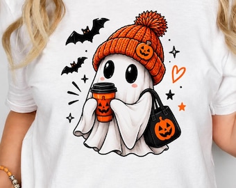 May include: A white t-shirt featuring a graphic of a white ghost wearing an orange knit beanie with a pumpkin patch. The ghost holds an orange coffee cup with a pumpkin face and a black tote bag with a pumpkin, surrounded by bats, stars, and a heart.