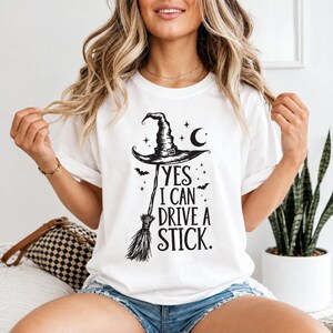 May include: A white short-sleeved t-shirt featuring a black graphic print of a witch hat atop a broomstick, accompanied by a crescent moon, stars, and bats. The text reads, "YES I CAN DRIVE A STICK." The design is centered on the chest area.