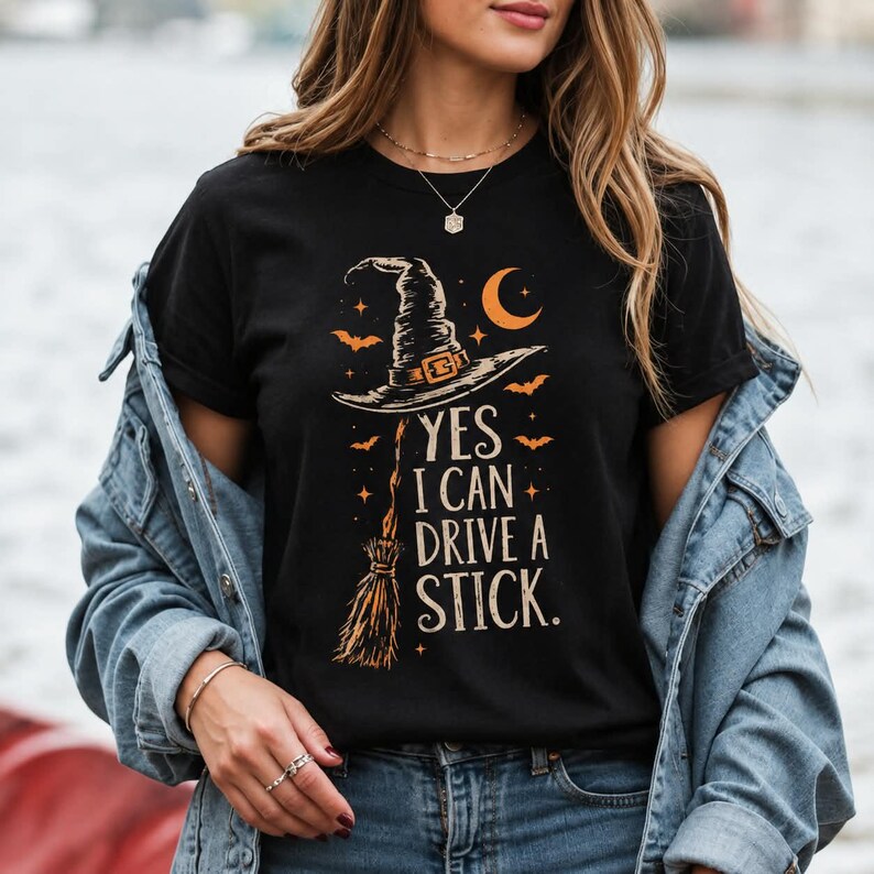 May include: A black t-shirt featuring a graphic of a witch hat and a broomstick in orange and cream colors. The text reads, YES I CAN DRIVE A STICK. The design includes small orange stars, bats, and a crescent moon surrounding the central illustration.