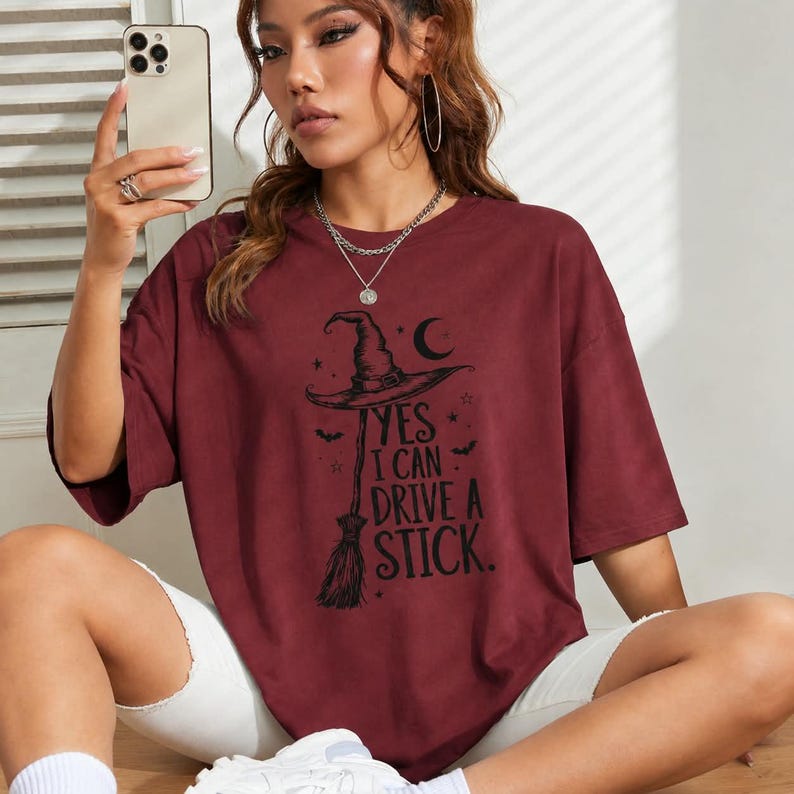 May include: A maroon short-sleeved t-shirt featuring a black graphic print of a witch hat, a broomstick, stars, bats, and a crescent moon. The text reads, "YES I CAN DRIVE A STICK." The shirt has a crew neckline and a loose, relaxed fit design.