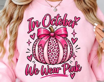 May include: A light pink crewneck sweatshirt featuring a graphic of a pumpkin with a pink leopard print pattern and a large pink bow. The text reads In October We Wear Pink, surrounded by small pink hearts and star-shaped accents on the front.