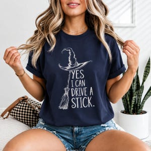 May include: A navy blue short-sleeve t-shirt featuring a white graphic of a witch hat and a broomstick. The text printed in white reads, YES I CAN DRIVE A STICK. The shirt has a crew neckline and is worn with blue denim shorts.