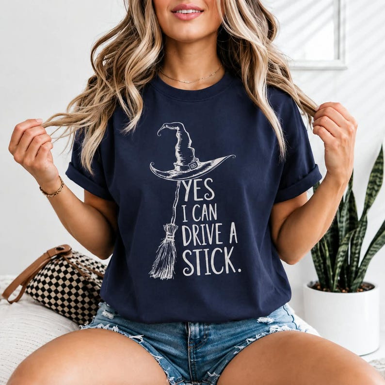 May include: A navy blue short-sleeve t-shirt featuring a white graphic of a witch hat and a broomstick. The text printed in white reads, YES I CAN DRIVE A STICK. The shirt has a crew neckline and is worn with blue denim shorts.