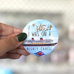 May include: A round pinback button featuring a light blue sky background with a graphic of a large cruise ship. The text reads I WISH I WAS ON A DISNEY CRUISE in black and gold lettering, with a small red Mickey Mouse icon above a blue wave graphic.