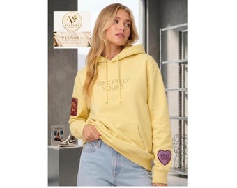 Puede incluir: Una sudadera con capucha de color amarillo pálido que presenta el texto SINCERELY YOURS impreso en un color claro en el pecho. La prenda incluye un bolsillo tipo canguro, un parche rectangular en la manga derecha y un parche morado en forma de corazón en la manga izquierda.