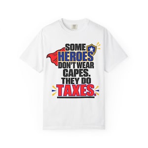 Könnte beinhalten: Ein weißes kurzärmeliges T-Shirt mit einem Grafikdruck, der den Text SOME HEROES DON'T WEAR CAPES. THEY DO TAXES. zeigt. Das Design umfasst ein rotes Cape, einen blau-gelben Schild mit einem weißen Stern sowie kräftige blaue, schwarze und rote Schrift.