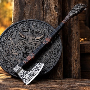 May include: A dark wooden axe with a silver-toned metal blade featuring intricate knotwork engravings and a wolf-head pommel, resting against a circular wooden shield embossed with a central wolf face and decorative patterns.
