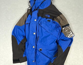 May include: A blue and black hooded jacket featuring The North Face logo on the chest, a front zipper with metal snap buttons, multiple pockets, adjustable drawstrings at the hood and hem, and a clear plastic utility sleeve on the left arm.