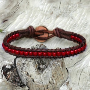 May include: A handcrafted bracelet featuring a braided brown leather band with a row of vibrant red beads. The bracelet is secured with a leather loop and a copper button clasp, showcasing a rustic and earthy aesthetic.