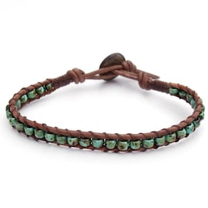 May include: A wrap bracelet featuring turquoise-colored beads and brown leather. The bracelet has a button closure and is designed to wrap around the wrist. The beads are evenly spaced along the leather band, creating a simple yet stylish accessory.