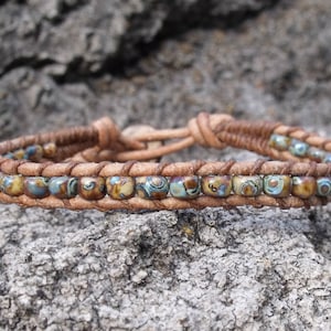 May include: Handmade bracelet featuring a braided leather band with a series of small, square, patterned beads in shades of blue, brown, and tan. The bracelet is secured with a leather knot closure.