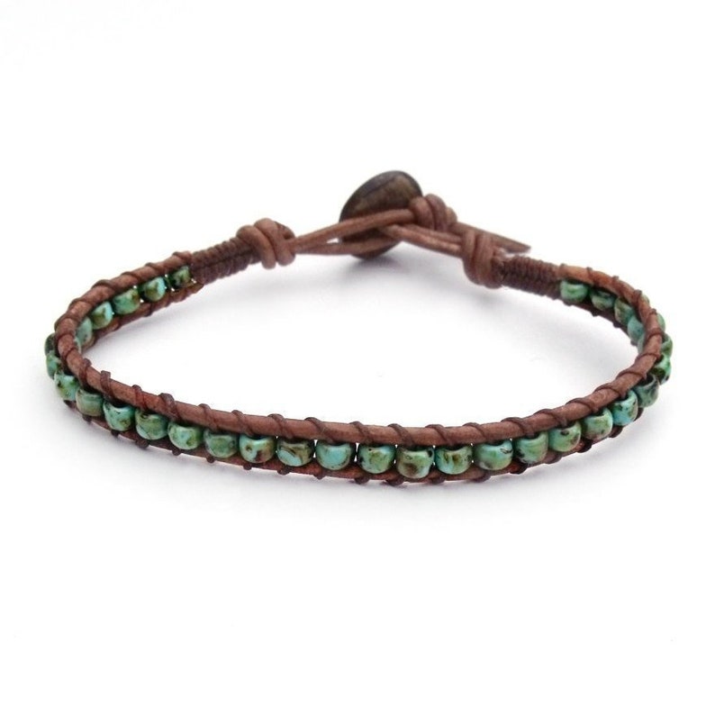 May include: A wrap bracelet featuring turquoise-colored beads and brown leather. The bracelet has a button closure and is designed to wrap around the wrist. The beads are evenly spaced along the leather band, creating a simple yet stylish accessory.