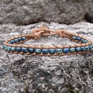 May include: A wrap bracelet featuring blue and green beads strung on a tan leather cord. The bracelet is secured with a button closure. The beads are a mix of solid and patterned, adding visual interest to the accessory.