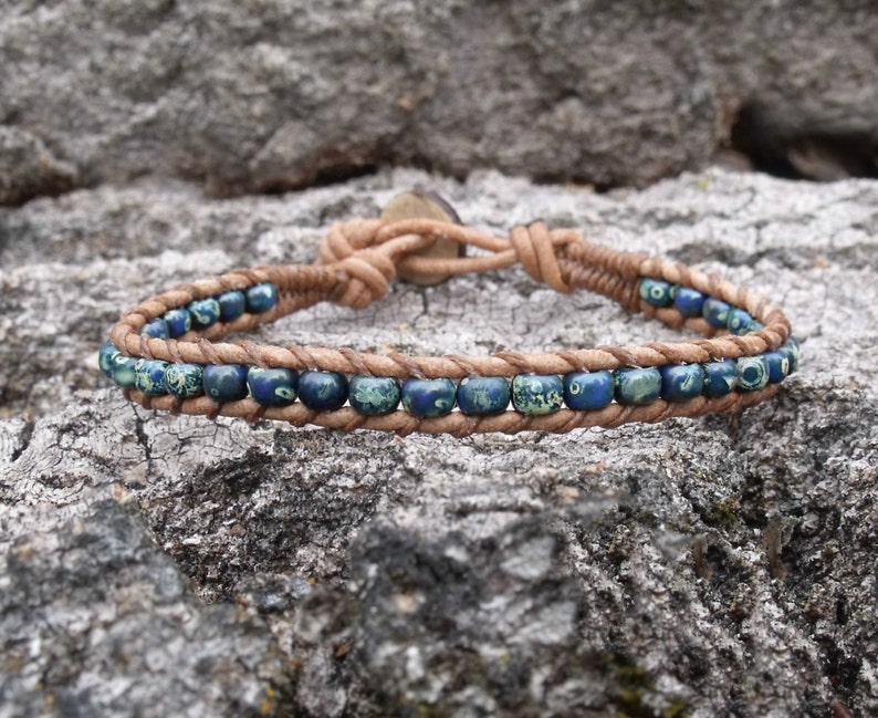 May include: A wrap bracelet featuring blue and green beads strung on a tan leather cord. The bracelet is secured with a button closure. The beads are a mix of solid and patterned, adding visual interest to the accessory.