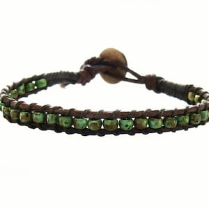 May include: A wrap bracelet featuring two rows of small, round, green beads. The beads are strung on a dark brown leather cord, with a matching leather strap and a wooden button closure. The bracelet is designed to wrap around the wrist.