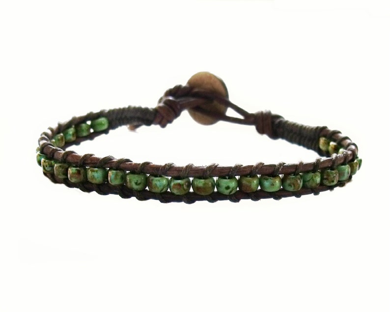 May include: A wrap bracelet featuring two rows of small, round, green beads. The beads are strung on a dark brown leather cord, with a matching leather strap and a wooden button closure. The bracelet is designed to wrap around the wrist.