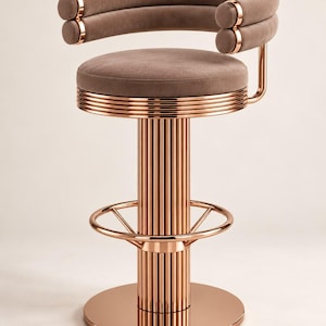 May include: A bar stool featuring a circular seat and a curved backrest upholstered in a taupe fabric. The frame is constructed from polished rose gold metal, showcasing a fluted cylindrical base, a circular footrest, and a flat round base.