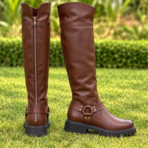 May include: A pair of brown knee-high boots featuring a smooth finish, a vertical silver-toned zipper running up the back of the shaft, and a decorative harness strap with metal hardware around the ankle, set on a thick black lug sole.