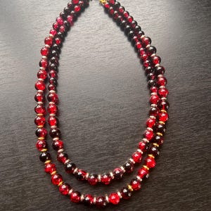 May include: A double-strand necklace featuring round, translucent red glass beads with a crackled texture. The beads are separated by small, metallic gold-toned spacers. The necklace is secured with a gold-toned circular clasp at the top center.