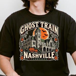 May include: A black t-shirt featuring a graphic print of a large stone building with a clock tower, an orange moon, bats, and two jack-o'-lanterns. The text reads GHOST TRAIN NASHVILLE TENNESSEE in a decorative font, surrounded by autumn leaves.