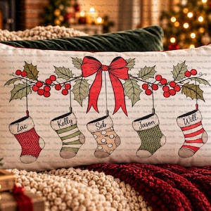 Op de afbeelding: Een rechthoekig sierkussen met een witte achtergrond en een feestelijke illustratie van vijf hangende kerstsokken. Elke sok heeft een uniek patroon en een naam: Zac, Kelly, Seb, Jason en Will. Een rode strik en hulsttakken zijn in het midden boven de sokken afgebeeld.