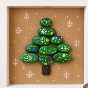 Peut inclure: Une décoration murale encadrée représentant un sapin de Noël composé de pierres peintes en vert disposées en triangle. Les pierres sont ornées de flocons de neige blancs et de petits points colorés. Le fond est beige avec des flocons blancs.